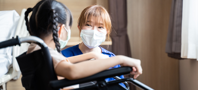 masked nurse speaking to a masked girl in a wheelchair